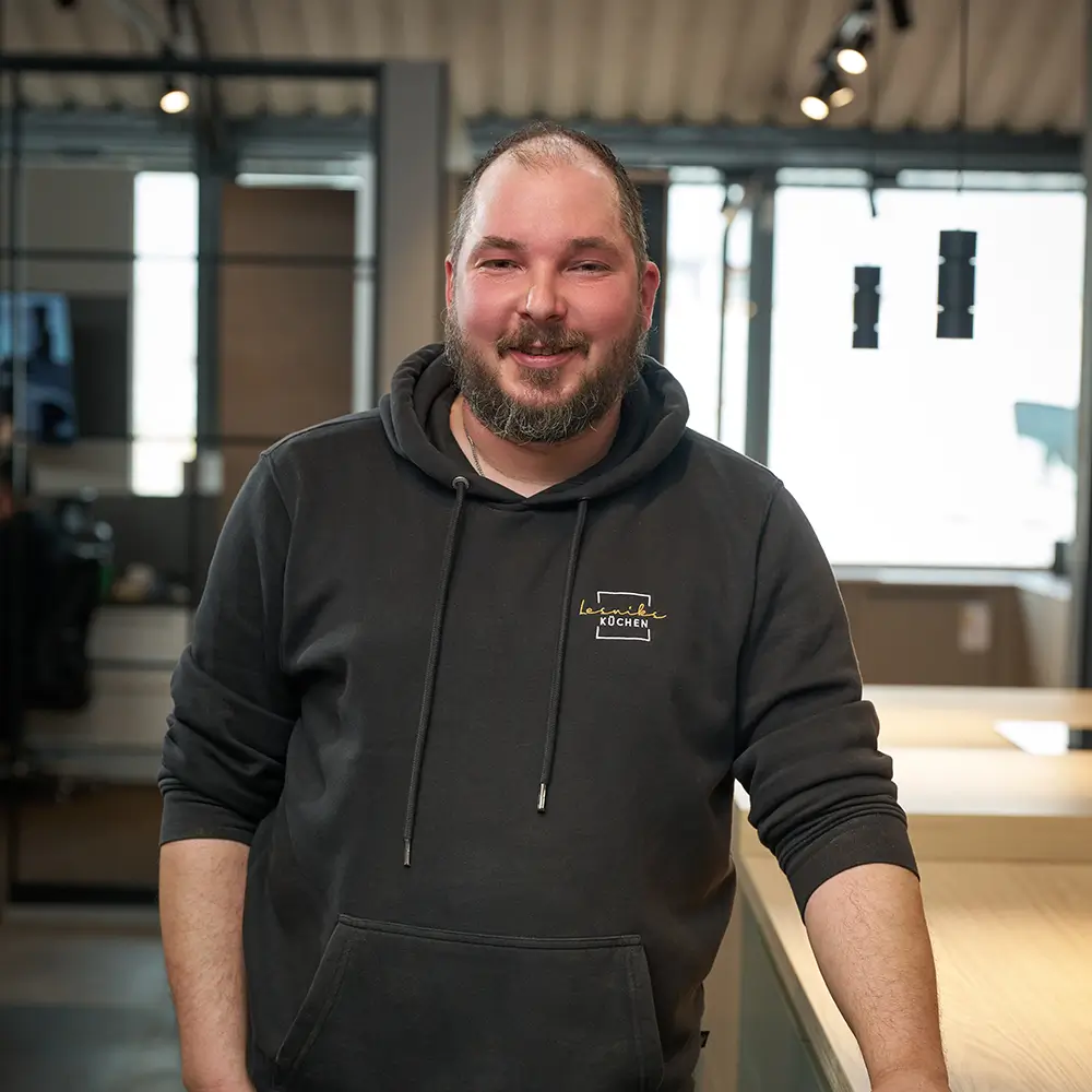 Eine lächelnde Person in dunklem Hoodie steht in einem modernen, warm beleuchteten Innenraum.