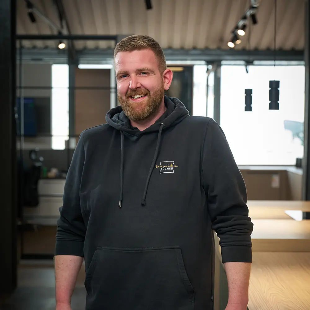 Eine lächelnde Person im Hoodie steht in einem modernen Büro mit neutralen Farben und offener Gestaltung.
