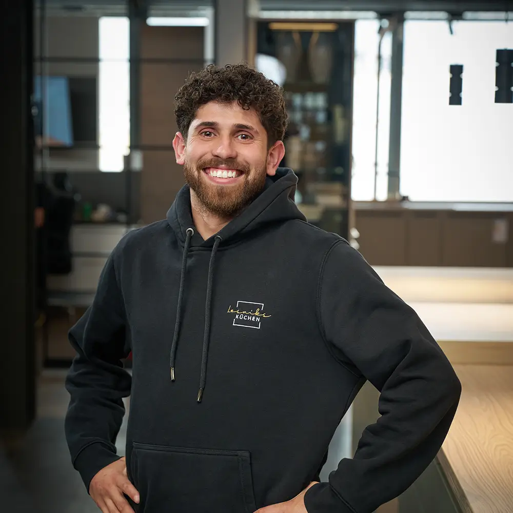 Eine lächelnde Person in einem schwarzen Hoodie steht in einem modernen, gut beleuchteten Raum.
