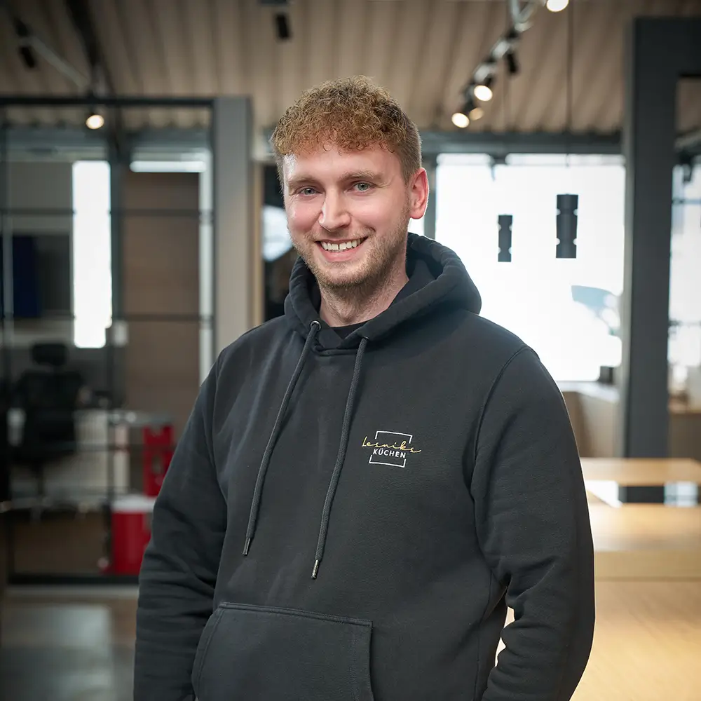 Lächelnde Person in dunkelgrauem Kapuzenpullover steht in modernem, lichtdurchflutetem Innenraum.