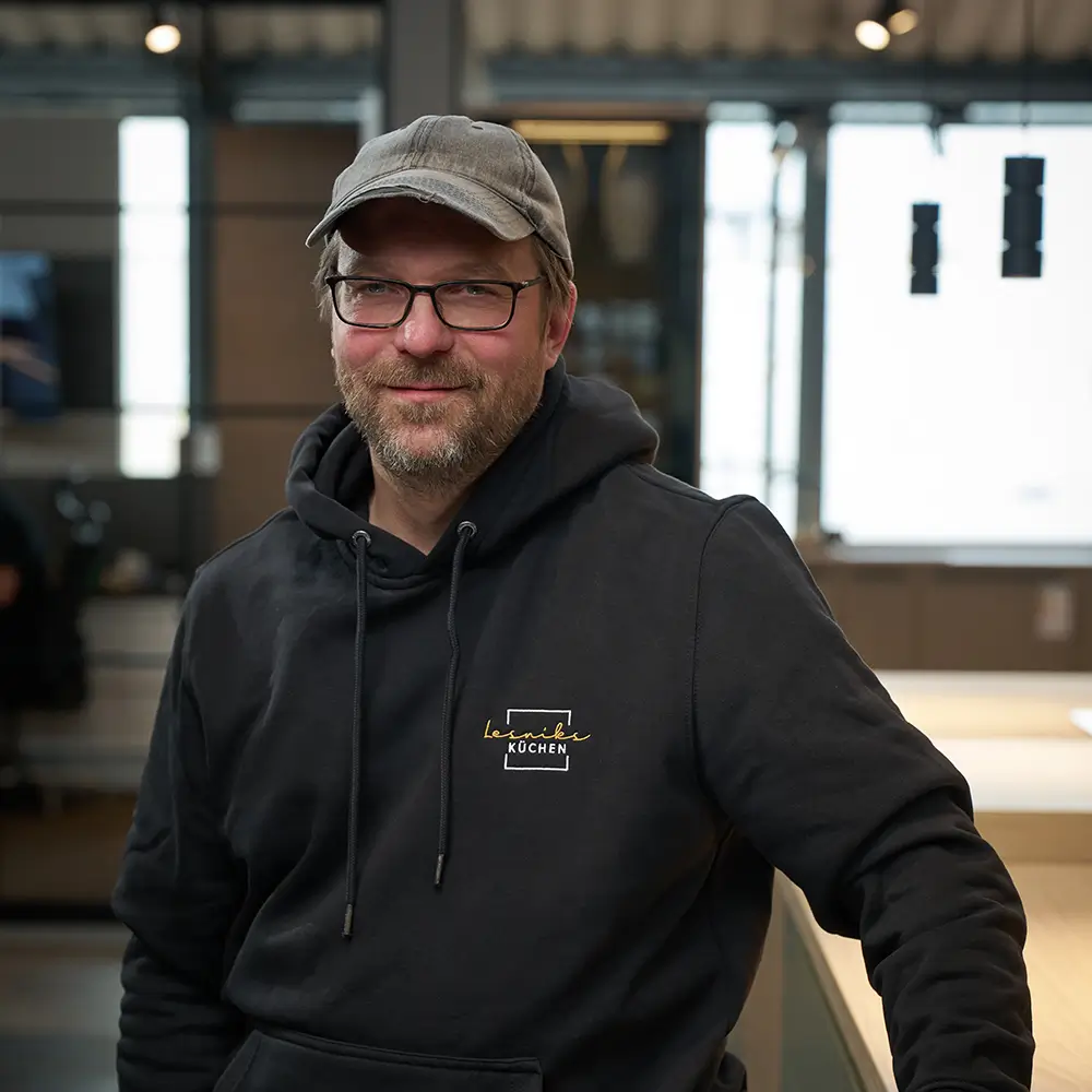 Ein Mann mit Brille und Kappe lächelt in einem modernen, lichtdurchfluteten Küchenraum.