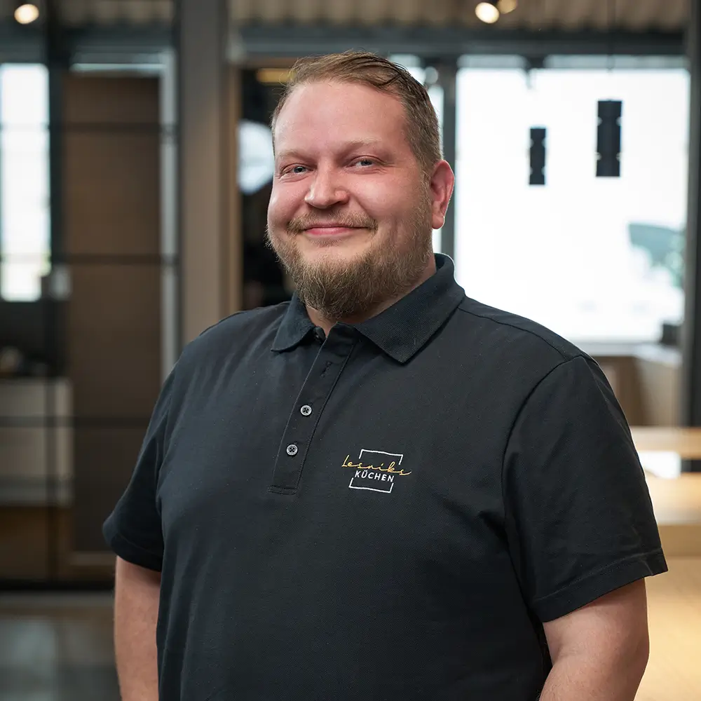 Eine Person in einem modernen Büro trägt ein schwarzes Polohemd mit Logo, wirkt freundlich und entspannt.