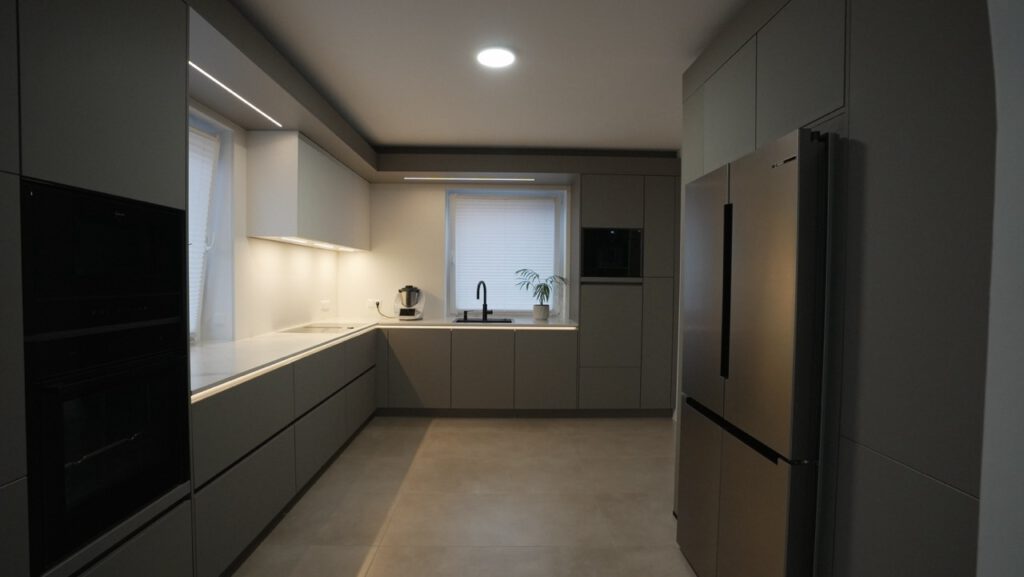 Moderne, minimalistische Küche in Grau und Weiß mit Kühlschrank, Herd, Spüle und warmem Licht.