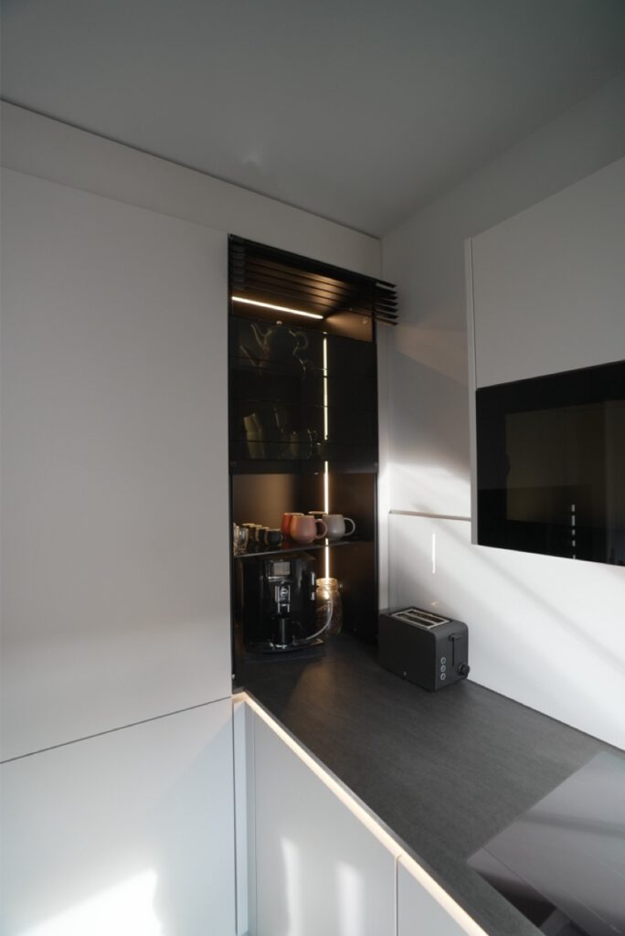 Moderne Küchenecke mit Kaffeemaschine, Toaster und Regal. Neutrale Töne, farbige Tassenakzente.