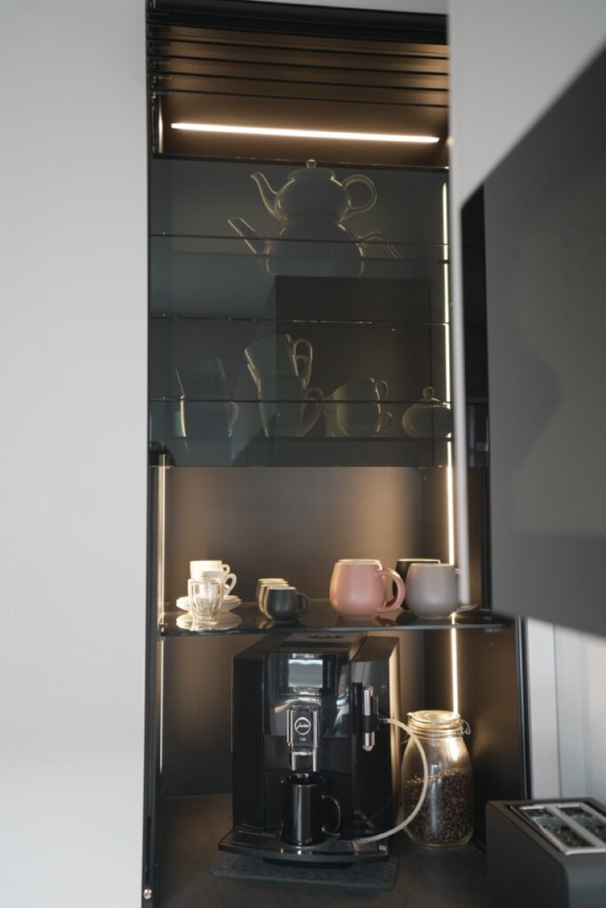 Moderne Küchenecke mit schwarzer Kaffeemaschine, Glasregalen und bunten Tassen in minimalistischer Anordnung.