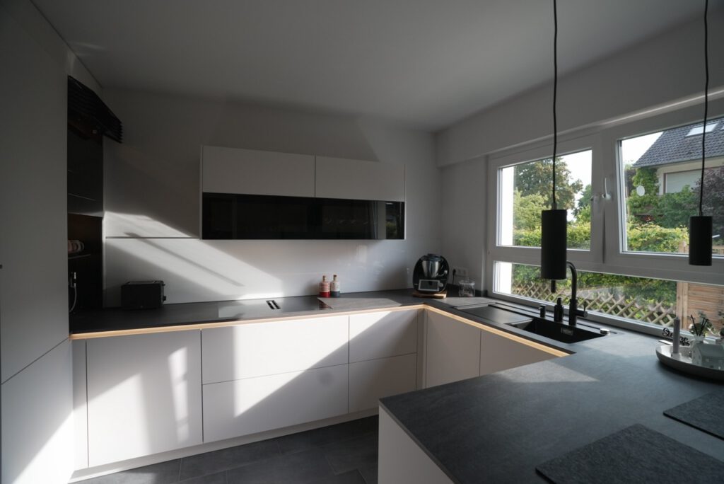 Moderne, aufgeräumte L-förmige Küche mit weißen Schränken, schwarzen Arbeitsflächen und natürlichem Licht.