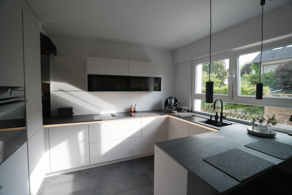 Moderne, minimalistische Küche mit weißen Schränken, dunkler Arbeitsplatte und großen Fenstern.