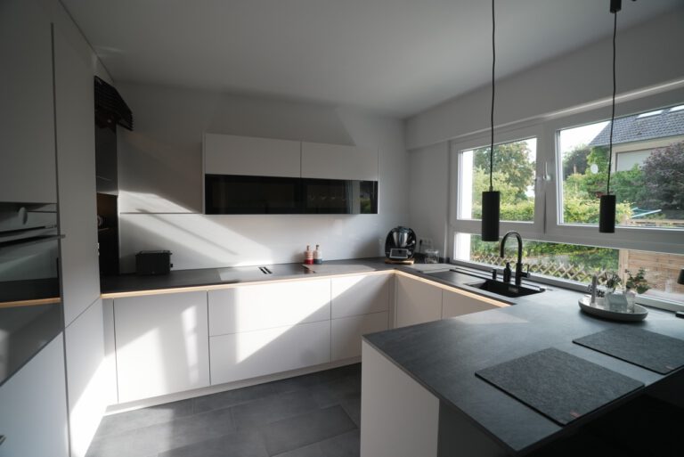 Moderne, minimalistische Küche mit weißen Schränken, dunkler Arbeitsplatte und großen Fenstern.