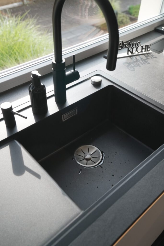 Moderne schwarze Küchenspüle mit Wasserhahn, Seifenspender und "Sterneküche"-Dekoration vor Fenster.