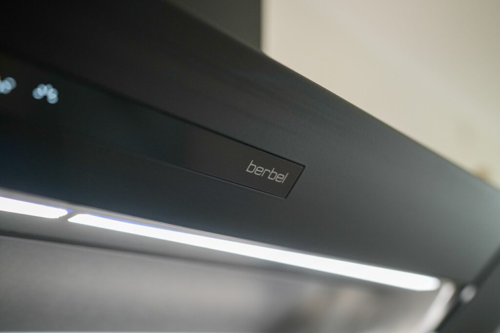 Ein Abschnitt einer modernen, dunklen Dunstabzugshaube mit "berbel"-Logo und heller LED-Beleuchtung.