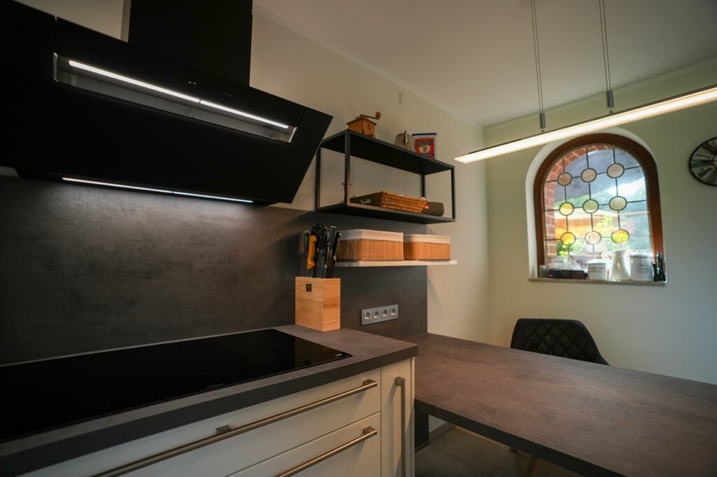 Moderne Küche mit schwarzem Induktionskochfeld, grauer Arbeitsfläche und buntem Rundfenster.