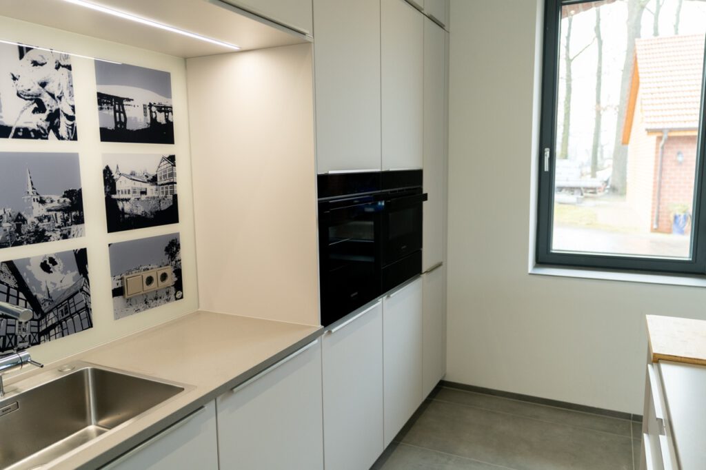 Moderne Küche mit Edelstahlspüle, weißen Schränken und Einbaugeräten, Blick auf Ziegeldach durch Fenster.