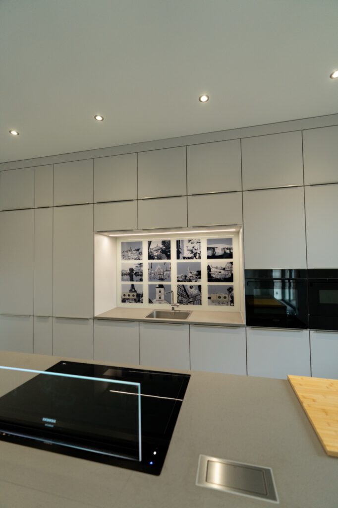 Moderne, minimalistische Küche in Weiß mit Induktionskochfeld, Edelstahlspüle und schwarzen Geräten.