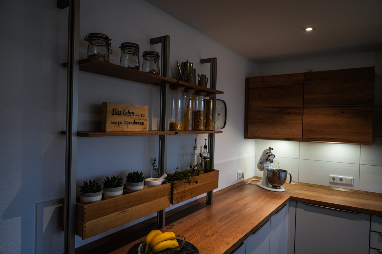 Moderne Küche mit Holzregalen, Vorratsgläsern, Pflanzen und einer glänzenden Küchenmaschine.