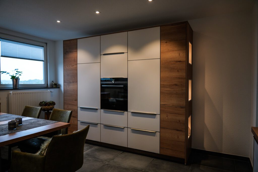 Moderne Küche mit weißen Schränken, Holzdetails, eingebautem Backofen und einem Esstisch mit grünen Stühlen.