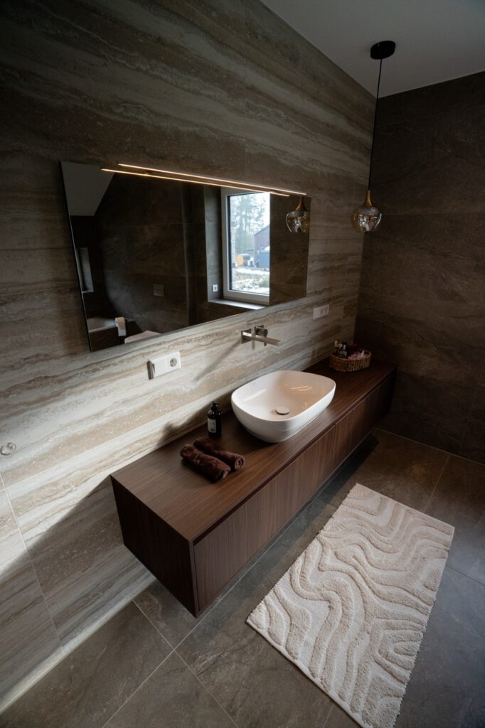 Modernes Badezimmer mit elegantem Design, Holzkonsole, beleuchtetem Spiegel und natürlichen Erdtönen.