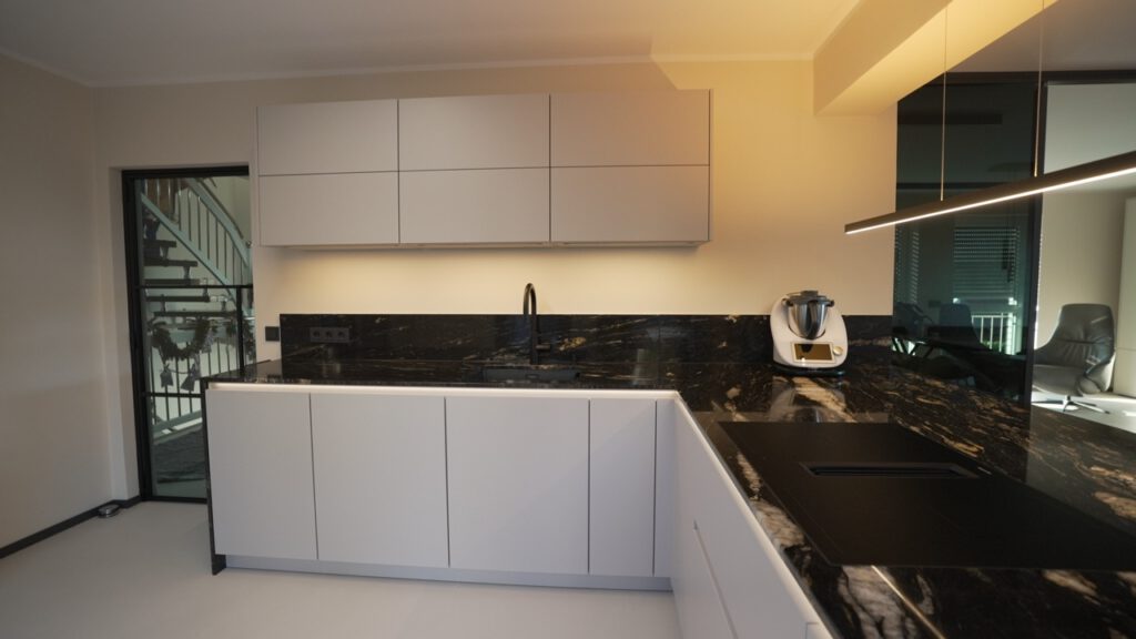Moderne, minimalistische Küche mit weißen Schränken, dunkler Arbeitsplatte und integriertem Kochfeld.