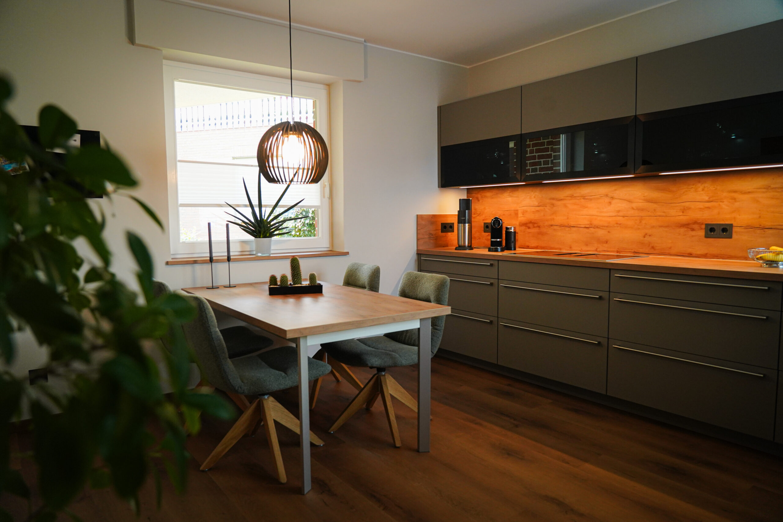 Moderne, aufgeräumte Küche mit Holztisch, grauen Schränken, Pflanzen und warmem Licht.