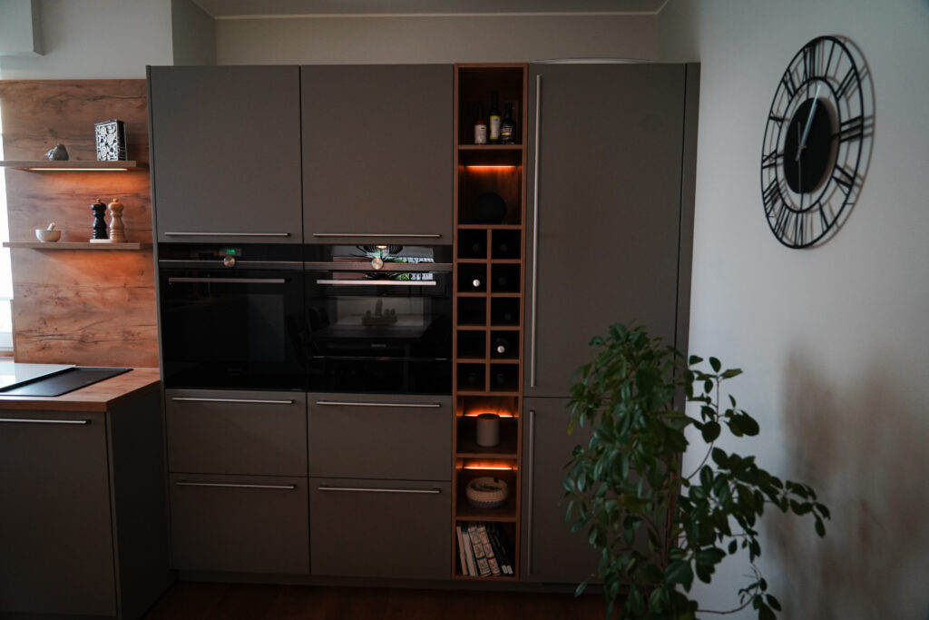 Moderne Küchenecke mit grauen Einbauschränken, offenes Regal, dekorative Uhr und Pflanze im Vordergrund.
