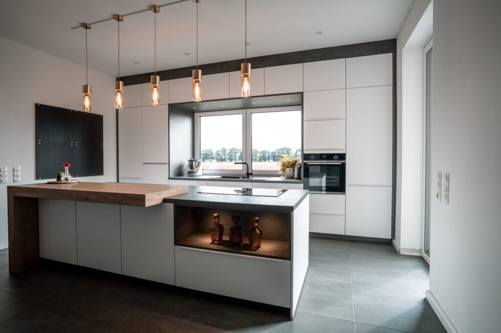 Moderne, minimalistische Küche mit Holzinsel, warmem Licht und großem Fenster mit Blick auf Bäume.