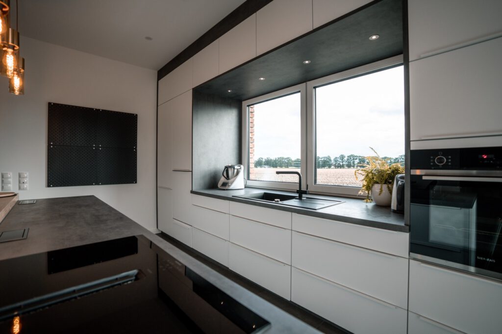 Moderne, minimalistische Küche mit weißen Schränken, schwarzer Arbeitsplatte und großem Fenster.