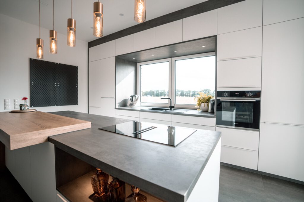 Moderne Küche mit weißem Design, dunkler Kücheninsel, viel natürlichem Licht und warmer Beleuchtung.