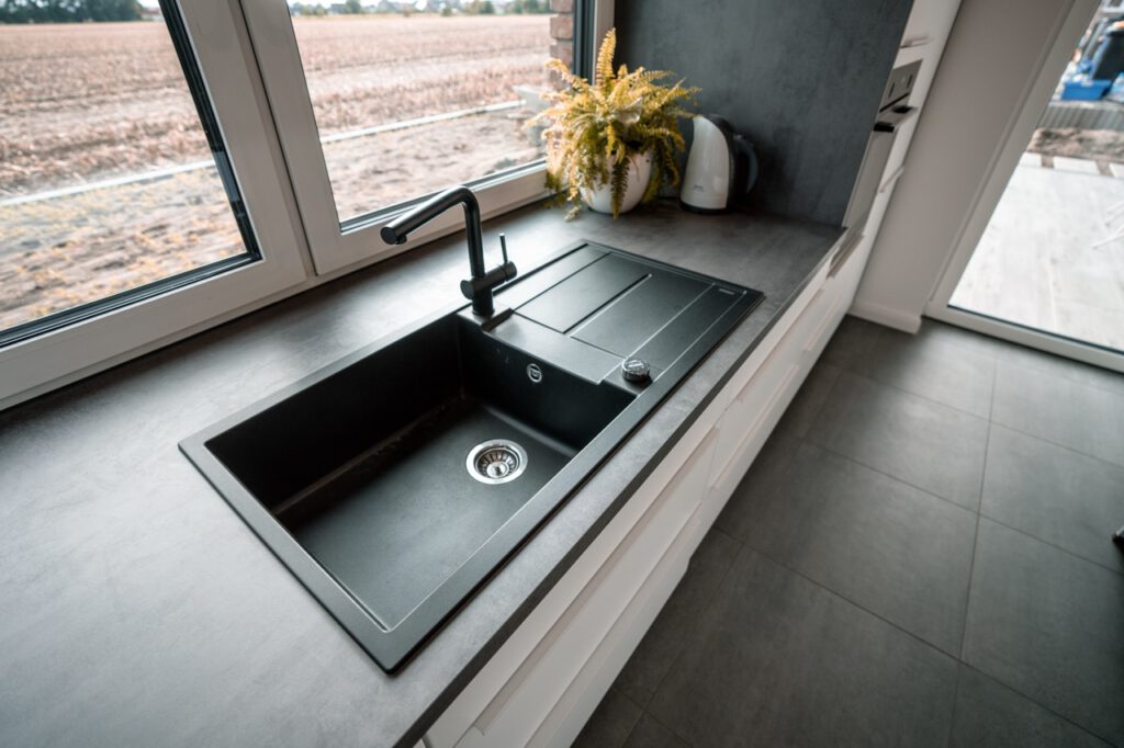 Moderne Küche mit schwarzer Spüle, Wasserkocher, Pflanze und großen Fenstern mit Blick ins Freie.