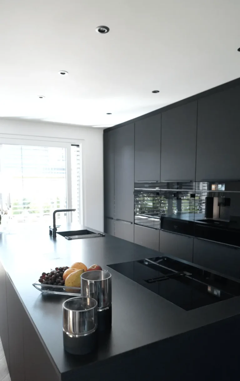 Moderne Küche mit dunkler Insel, glänzenden Schränken, Früchteschale und Kochfeld, elegant beleuchtet.