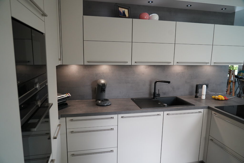 Moderne, minimalistische Küche mit weißen Schränken, grauer Arbeitsplatte und buntem Früchtekorb.
