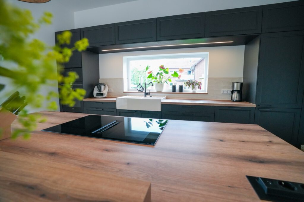 Moderne Küche mit schwarzer Schränken, Holzarbeitsplatte, Induktionskochfeld und Pflanzen am Fenster.