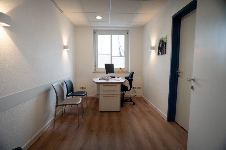 Ein kleiner, minimalistischer Büroraum mit Schreibtisch, Stühlen und einem Fenster, das Tageslicht hereinlässt.