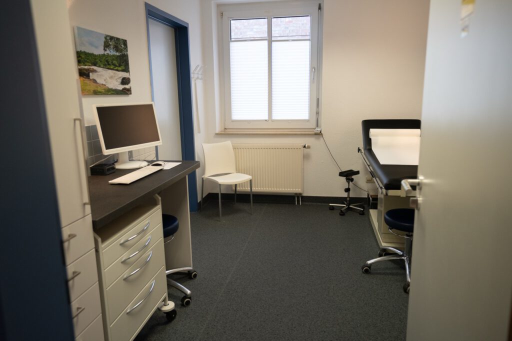 Ein leeres, funktionales Arztzimmer mit Schreibtisch, Monitor, Untersuchungsliege und weißen Wänden.