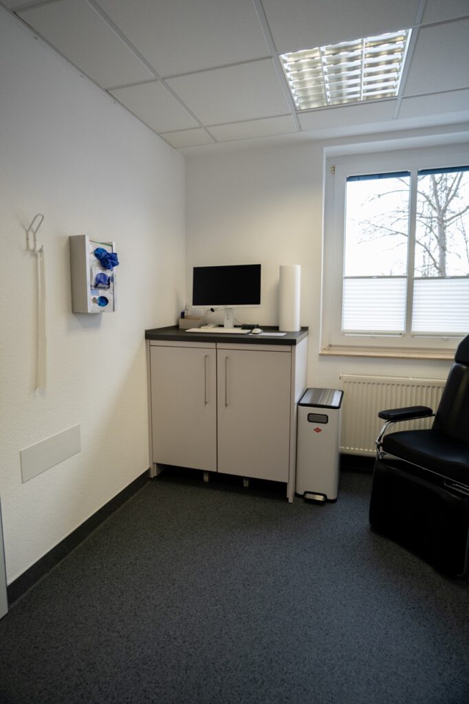 Kleines, sauberes Behandlungszimmer mit Schrank, Computer, Handschuhspender und schwarzem Stuhl.