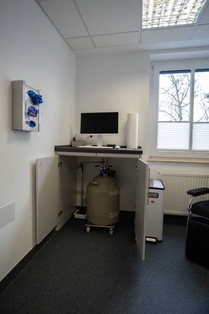 Ein kleiner, steriler Raum mit Computer, Kryotank und blauen Handschuhen an der Wand.