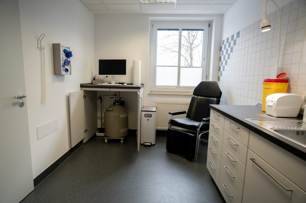 Ein leerer, steriler Untersuchungsraum mit Stuhl, Computer, Waschbecken und gelbem Abfallbehälter.