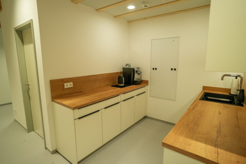 Moderne Büroküche mit Kaffeemaschine, Wasserkocher und Waschbecken auf heller Holzarbeitsplatte.