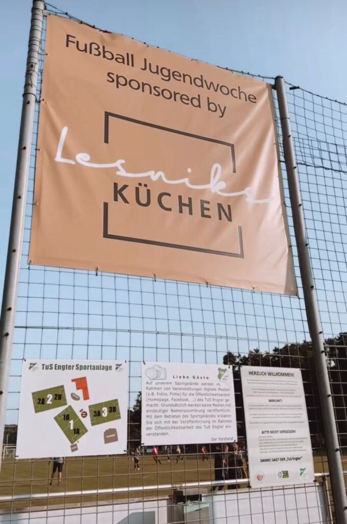 Banner "Fußball Jugendwoche" am Zaun, mit Lageplan und Infoschildern auf Sportgelände.