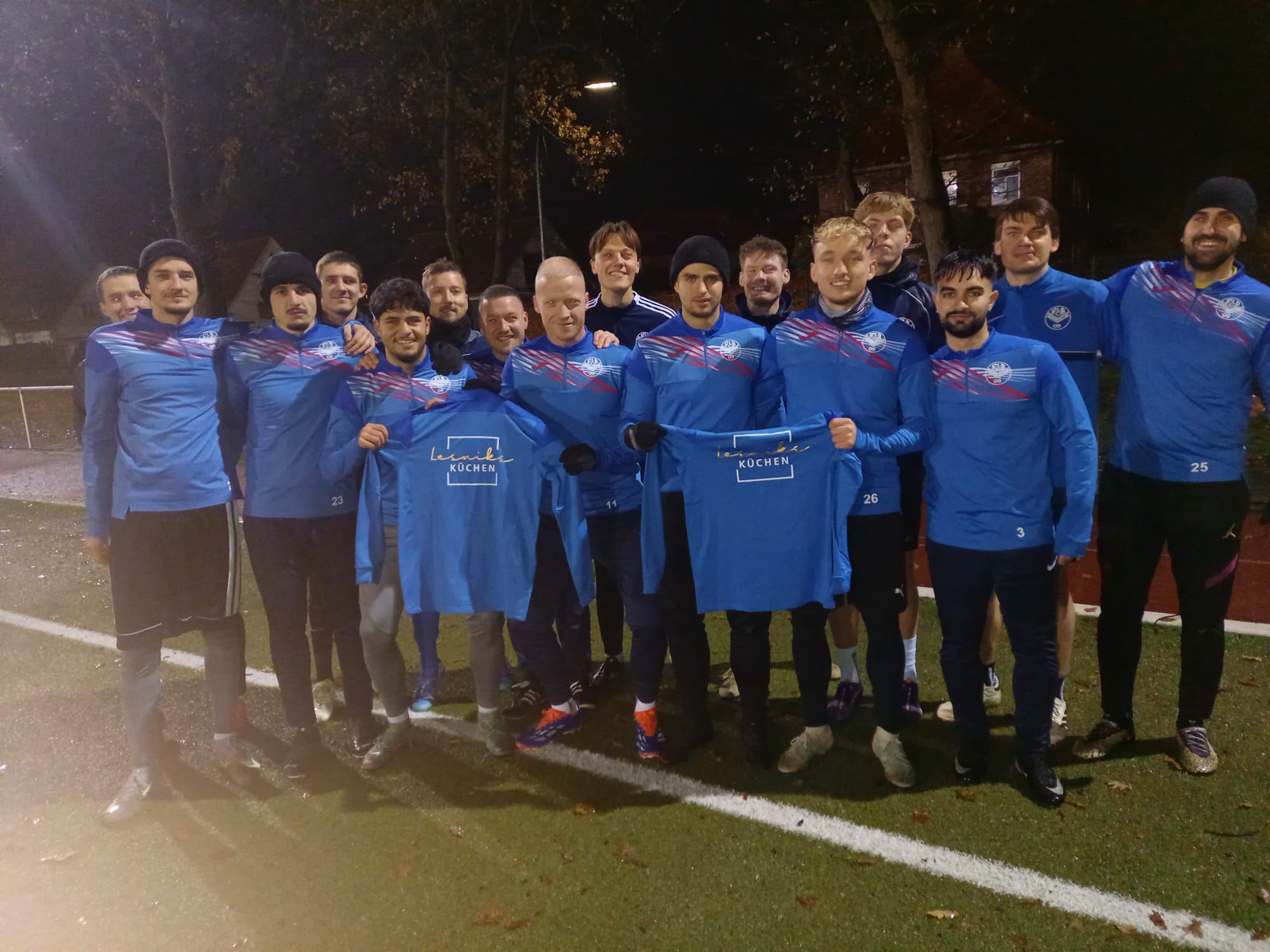 Eine Gruppe von 15 Personen in blauen Trainingsanzügen posiert lächelnd auf einem nächtlichen Fußballplatz.