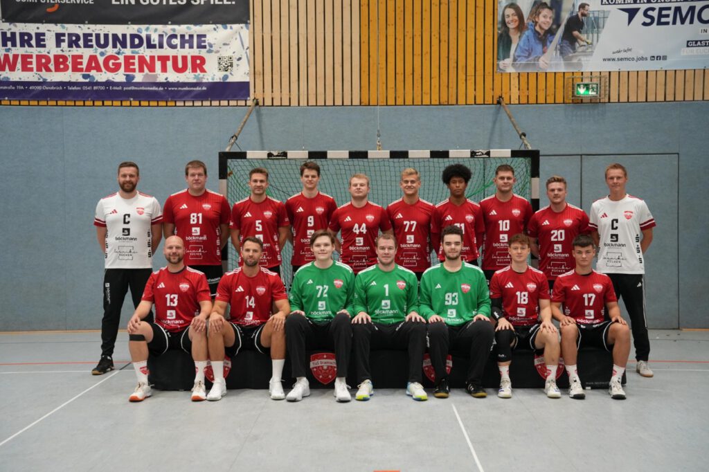 Ein Teamfoto einer Handballmannschaft in roten und grünen Trikots vor einem Handballtor in der Sporthalle.