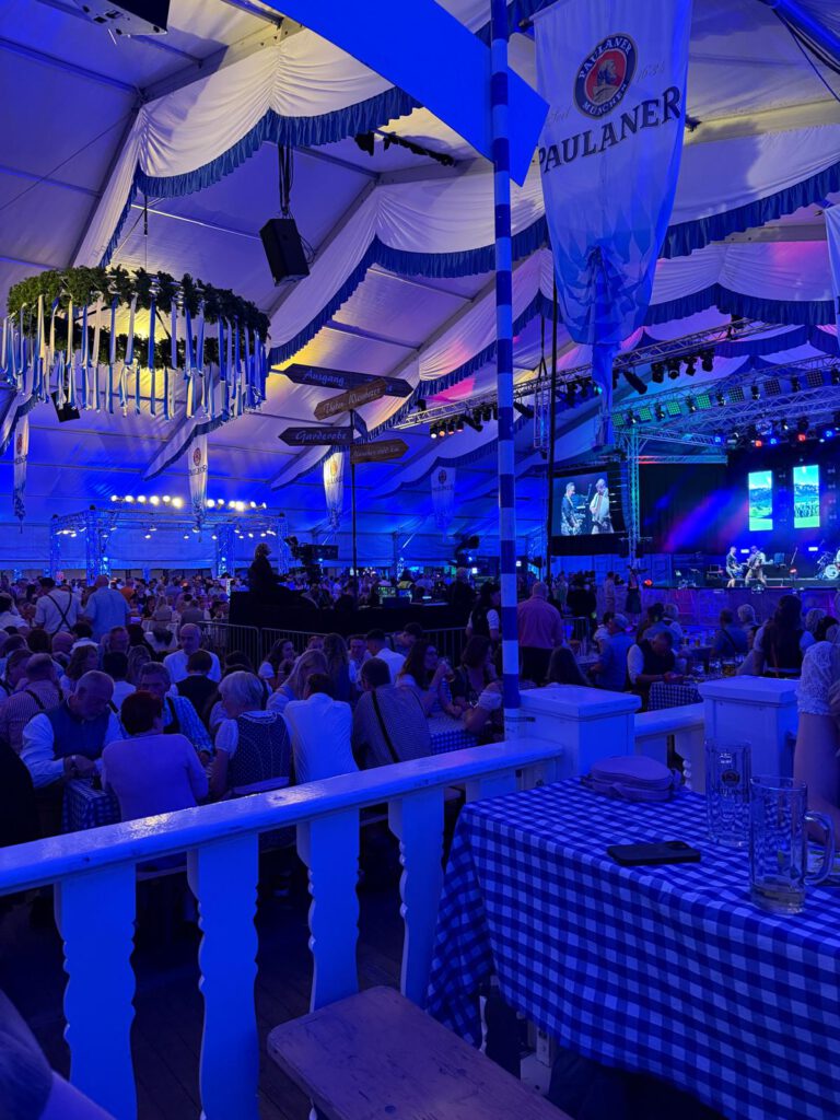 Festzelt mit Menschen an blau-weiß gedeckten Tischen, Musikbühne im Hintergrund, festliche Stimmung.