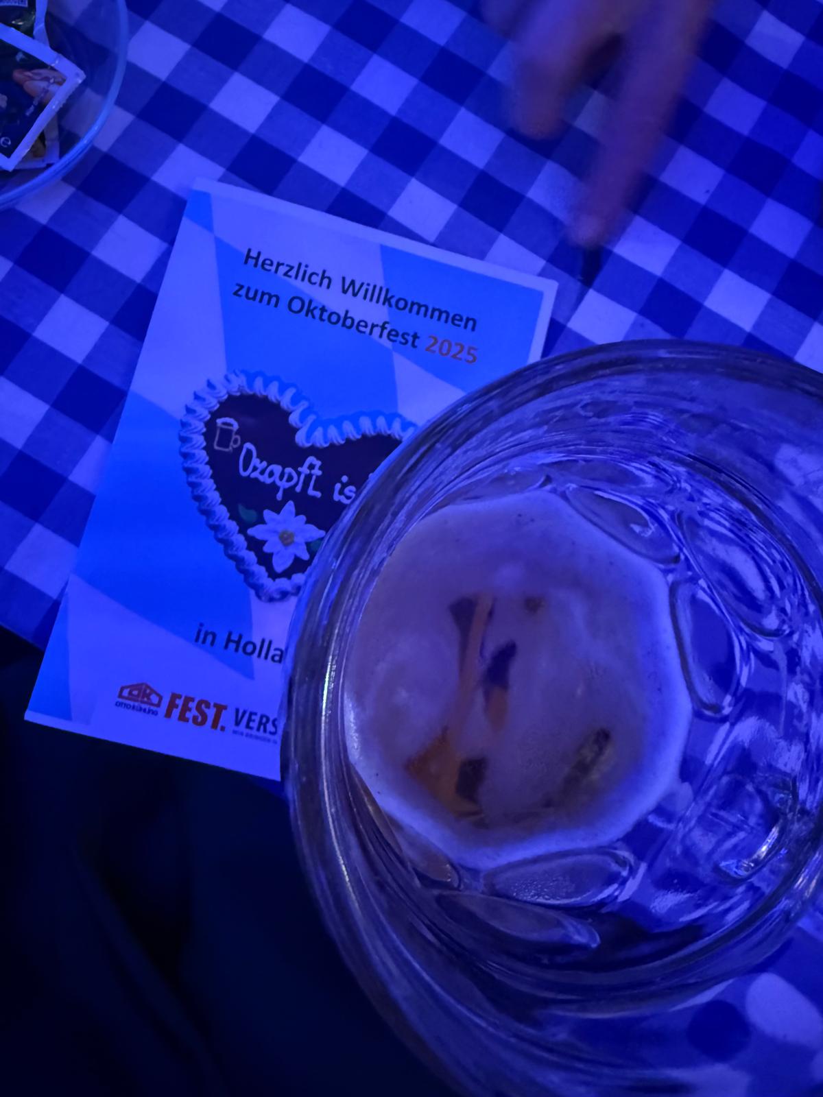 Ein Bierkrug und ein Oktoberfest-Flyer auf blau-weißer Tischdecke, Hand greift nach dem Flyer.