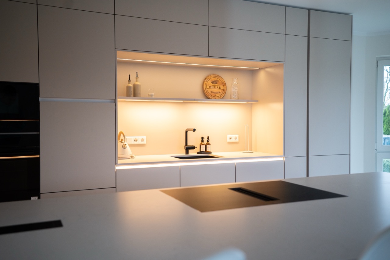 Moderne, minimalistische Küche mit hellen Fronten, schwarzer Armatur und dekorativ beleuchtetem Regal.