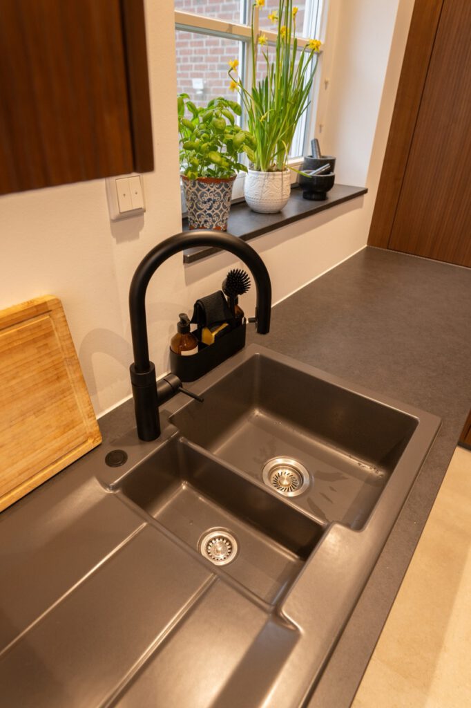 Moderne Küche mit grauer Spüle, schwarzem Wasserhahn, Pflanzen auf der Fensterbank und Holzschneidebrett.