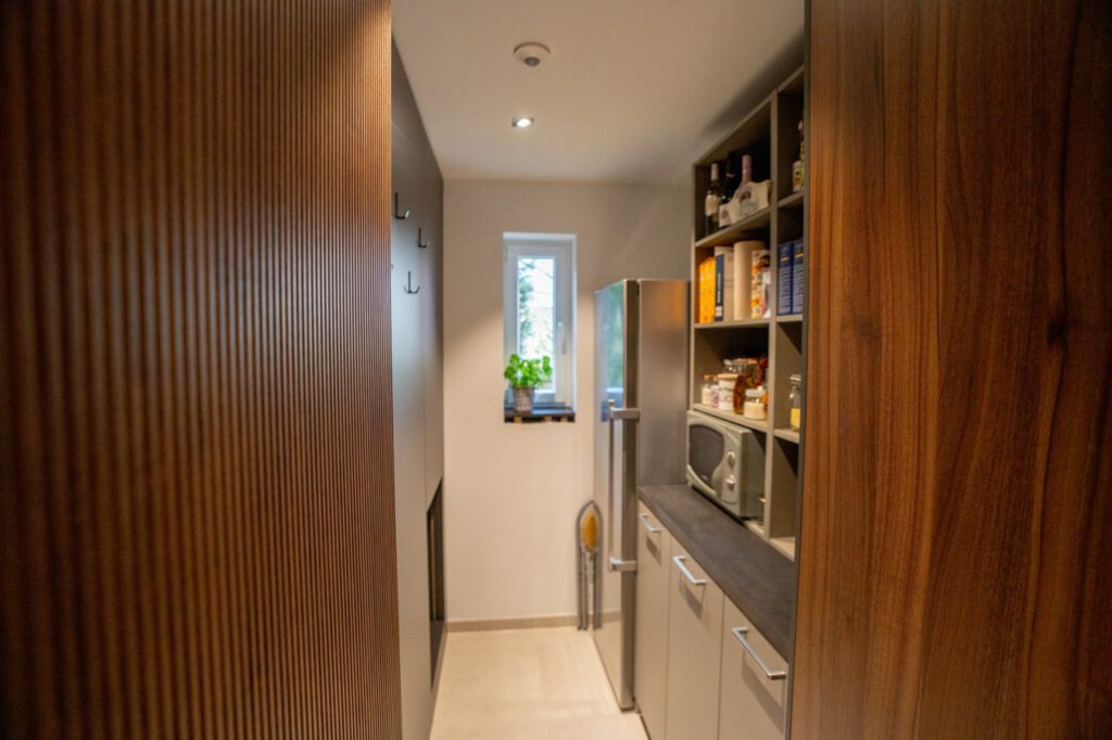 Moderne Speisekammer mit Kühlschrank, Regalen voller Lebensmittel und einer Mikrowelle, umgeben von Holz.