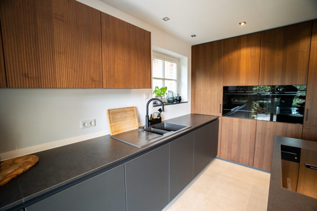 Moderne Küche mit warmen Holzschränken, dunkler Arbeitsplatte, Edelstahlspüle und Pflanzen am Fenster.