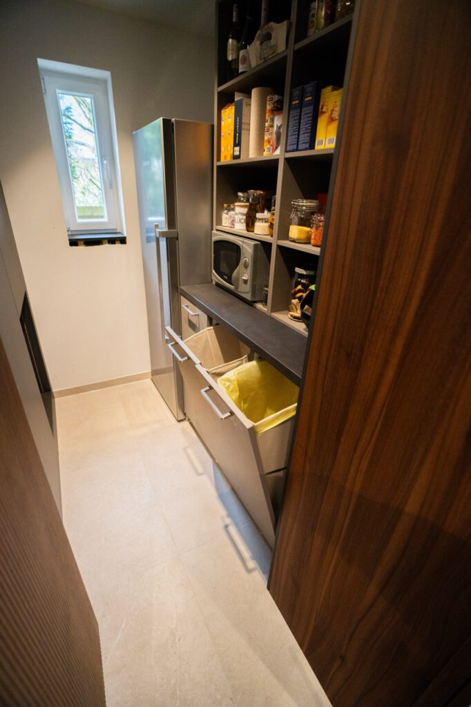 Ein aufgeräumter Lagerraum mit Kühlschrank, Regalen voller Lebensmittel und offenen Müllschubladen.