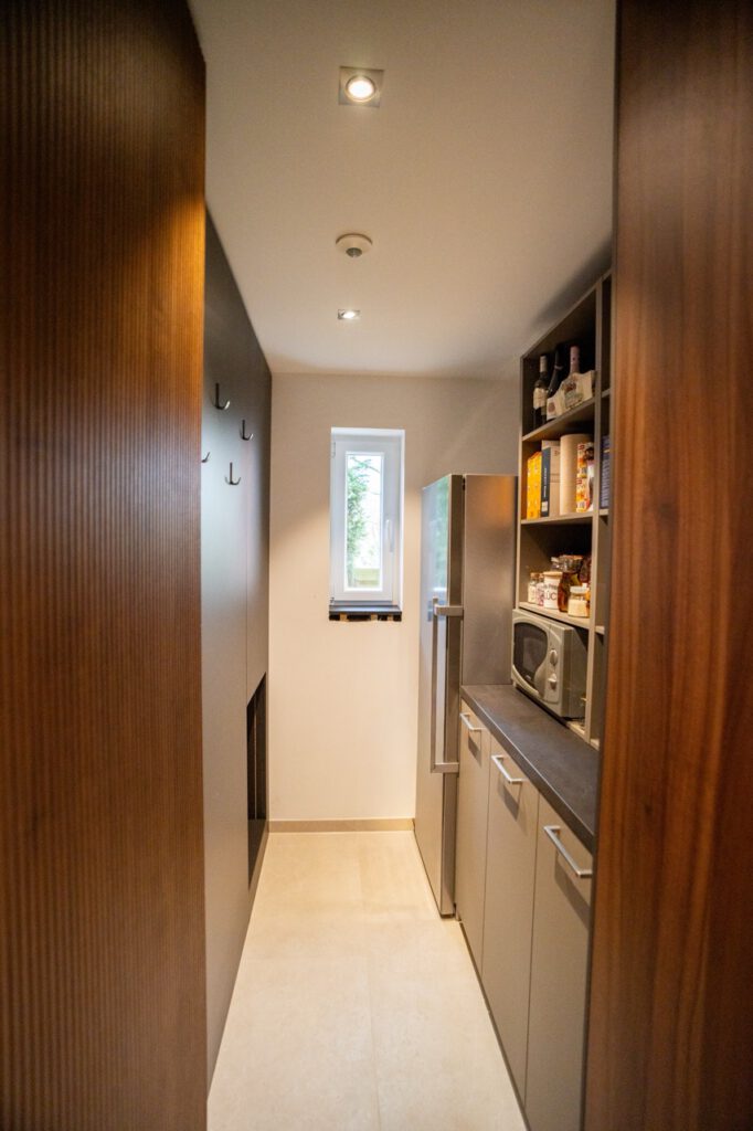 Ein schmaler Hauswirtschaftsraum mit modernen Schränken, Regalen und einem Kühlschrank.