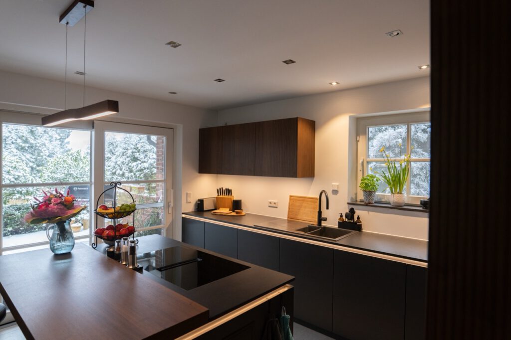 Moderne, aufgeräumte Küche mit dunkler Arbeitsplatte, Blumenstrauß, Obst und Blick auf verschneite Landschaft.