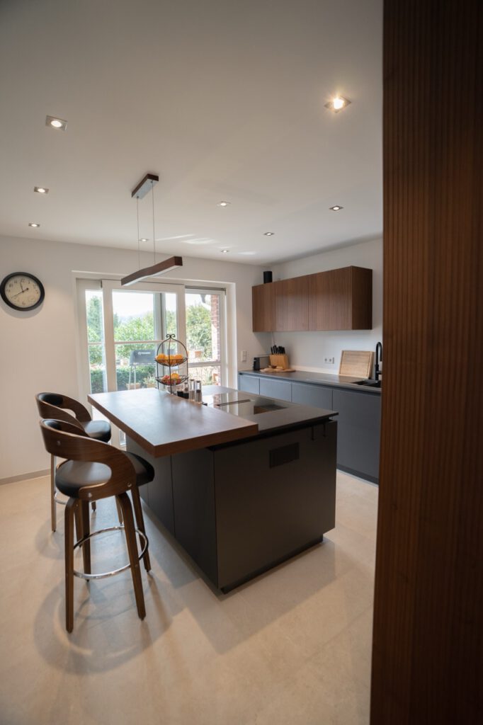 Moderne, minimalistische Küche mit dunkler Kücheninsel, Holzbarhockern und viel natürlichem Licht.
