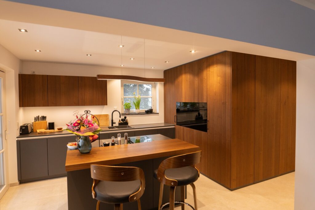 Moderne, aufgeräumte Küche mit Holz- und Grautönen, zentraler Insel und stilvollen Barhockern.